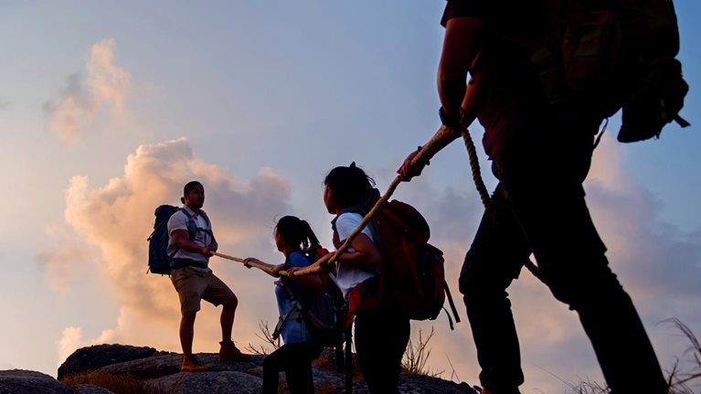 Group of friends hikers climbing up silhouette mountain cliff.