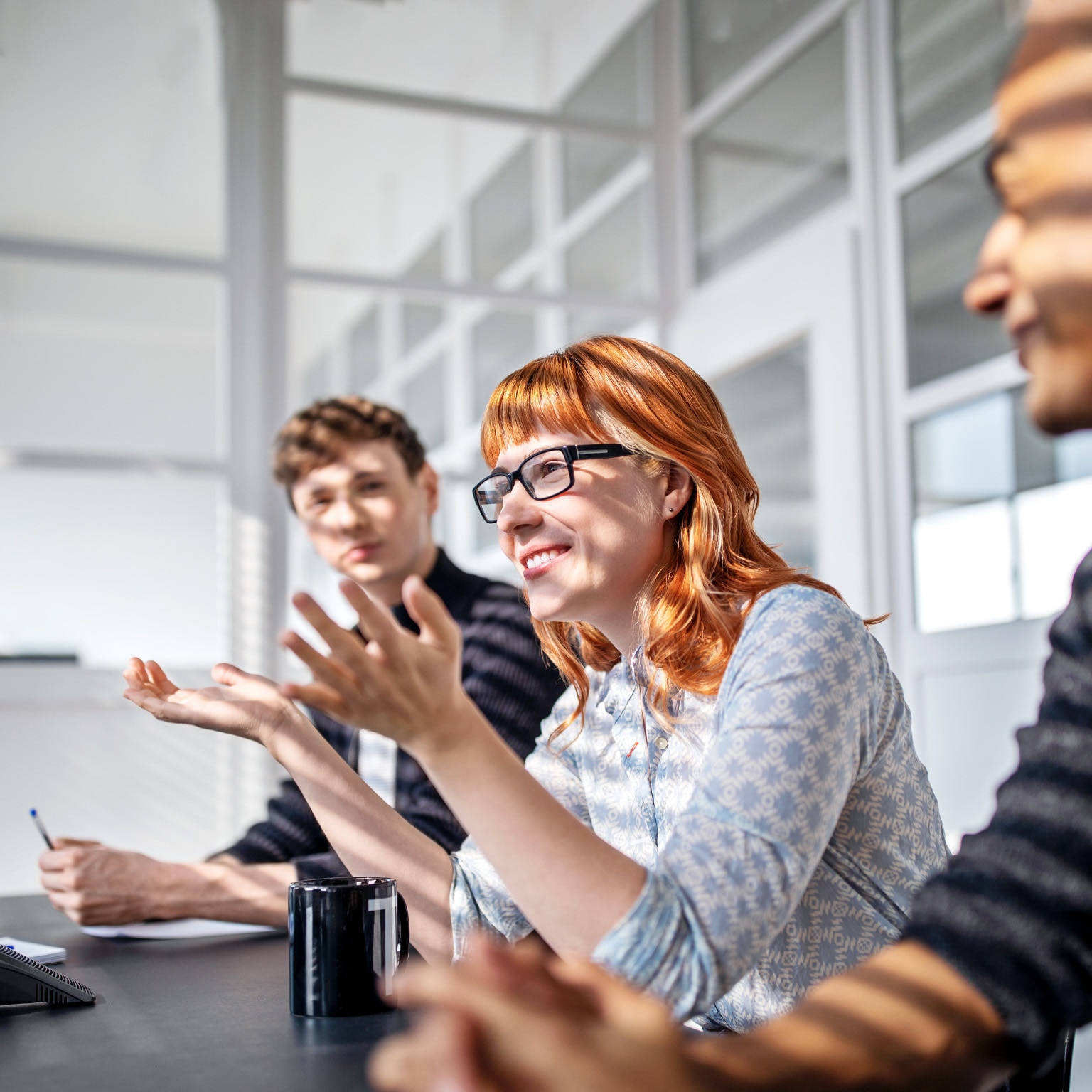 Three happy and engaged employees sitting at a board room table during a meeting. A woman sitting at the center of the group leans in to speak while gesturing expressively with her hands.  