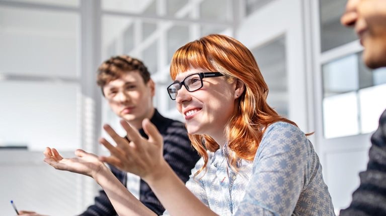 Three happy and engaged employees sitting at a board room table during a meeting. A woman sitting at the center of the group leans in to speak while gesturing expressively with her hands.