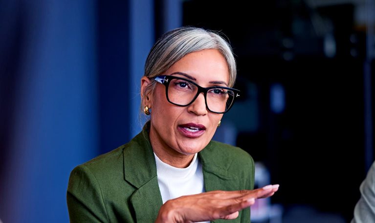 A mature businesswoman in a green blazer and glasses is speaking confidently while gesturing with her hands during a boardroom meeting.
