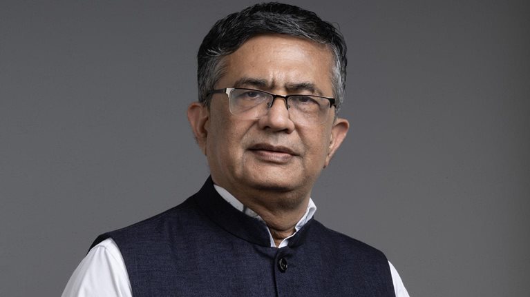 Ashishkumar Chauhan, with short hair and glasses, is wearing a white shirt and a dark blue Nehru vest, standing against a gray background.