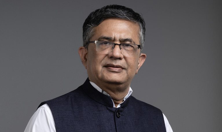 Ashishkumar Chauhan, with short hair and glasses, is wearing a white shirt and a dark blue Nehru vest, standing against a gray background.