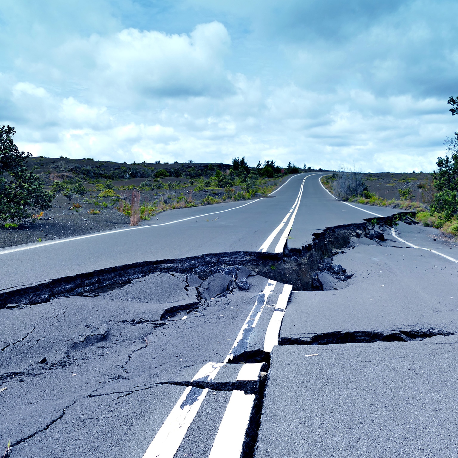 Image of a paved road that is broken and buckled with a huge crack running down its center. 