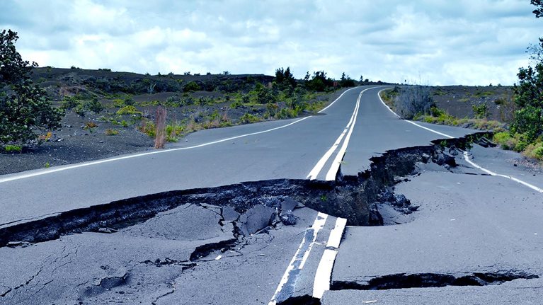 Image of a paved road that is broken and buckled with a huge crack running down its center.