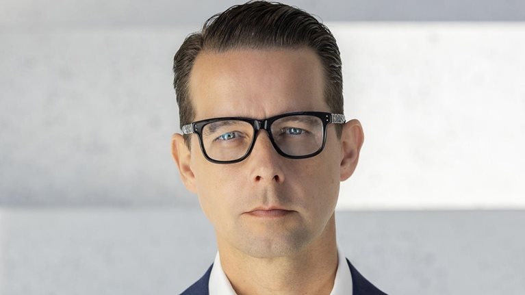 Jacob Aarup-Andersen is depicted in a portrait with a stern look, wearing glasses, a deep blue suit coat, and a white button-down shirt.