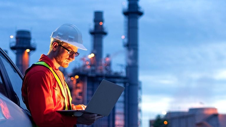 Engineer wearing safety helmet using laptop with oil refinery background