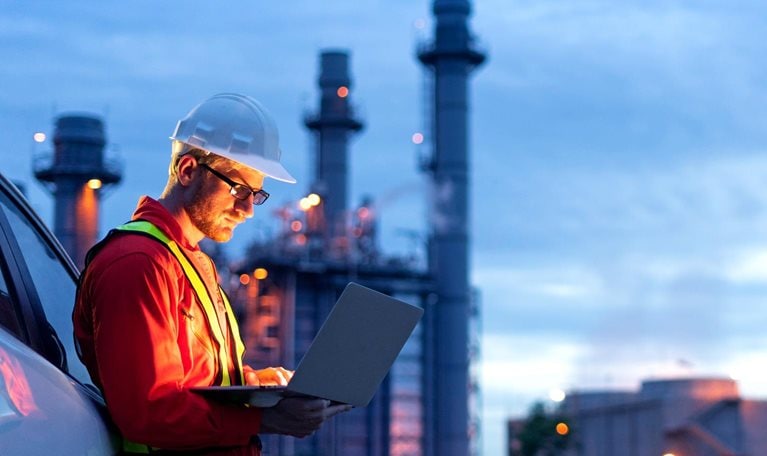 Engineer wearing safety helmet using laptop with oil refinery background