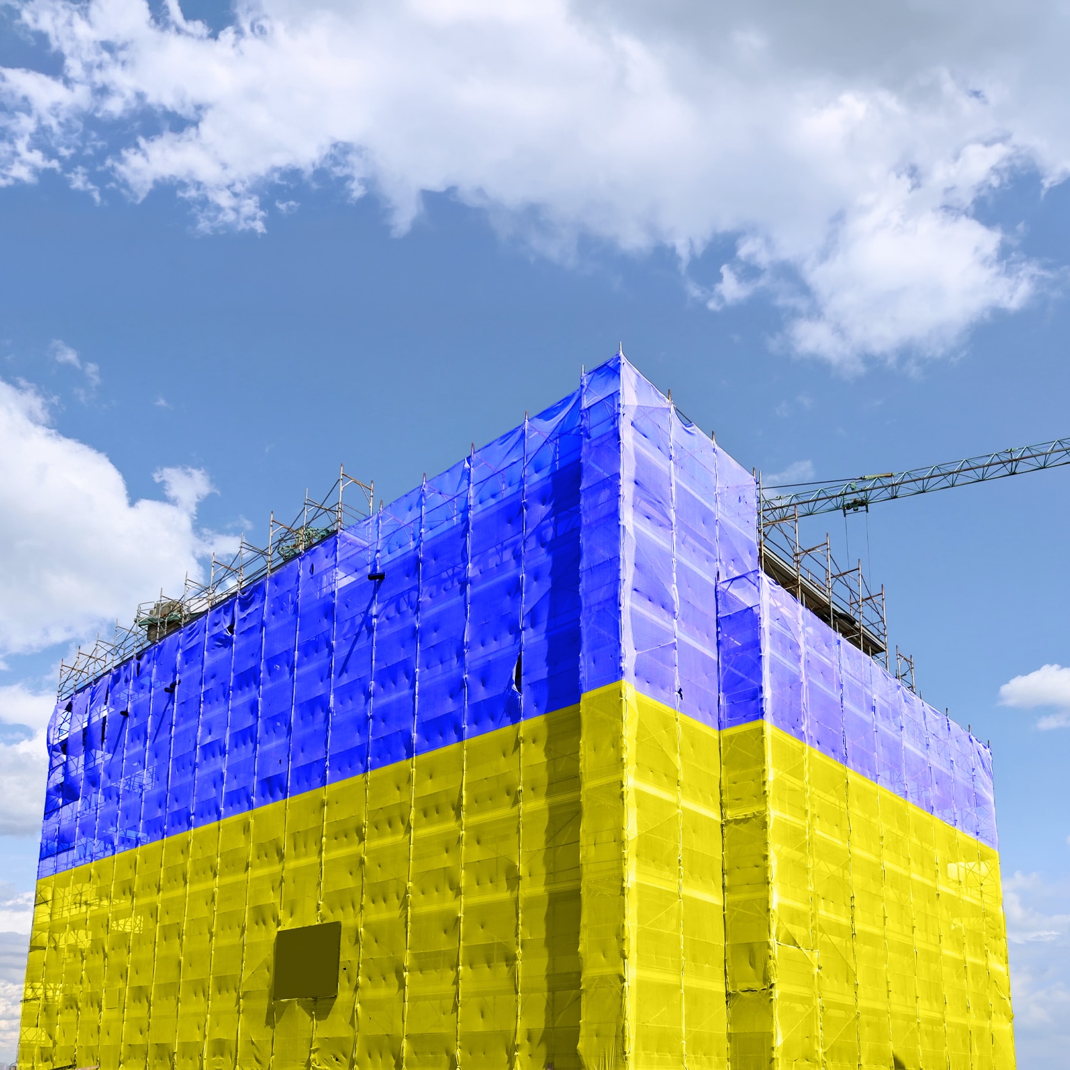 A building under construction is wrapped in scaffolding and covered with large sheets in bright blue and yellow, resembling the colors of the Ukrainian flag. A crane extends above the structure against a partly cloudy sky, emphasizing the ongoing construction work.