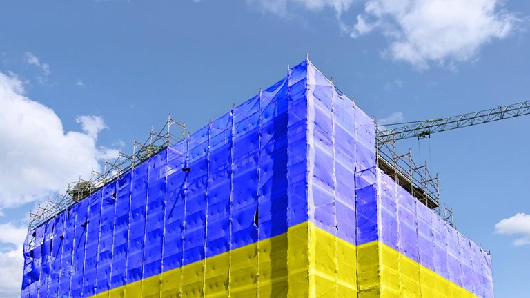 A building under construction is wrapped in scaffolding and covered with large sheets in bright blue and yellow, resembling the colors of the Ukrainian flag. A crane extends above the structure against a partly cloudy sky, emphasizing the ongoing construction work.