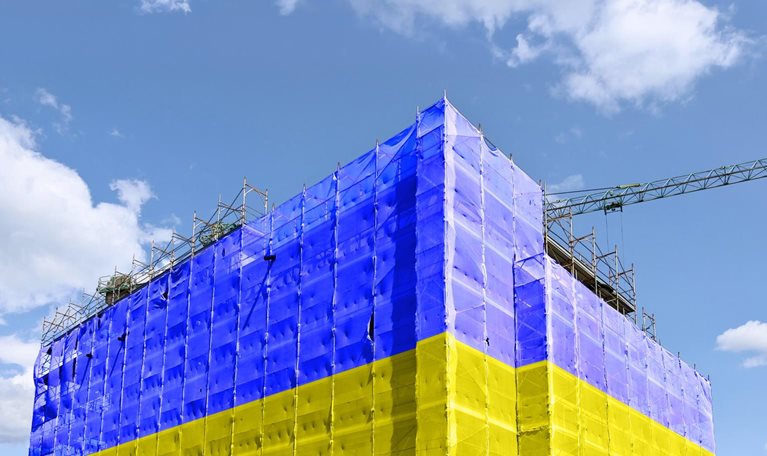 A building under construction is wrapped in scaffolding and covered with large sheets in bright blue and yellow, resembling the colors of the Ukrainian flag. A crane extends above the structure against a partly cloudy sky, emphasizing the ongoing construction work.