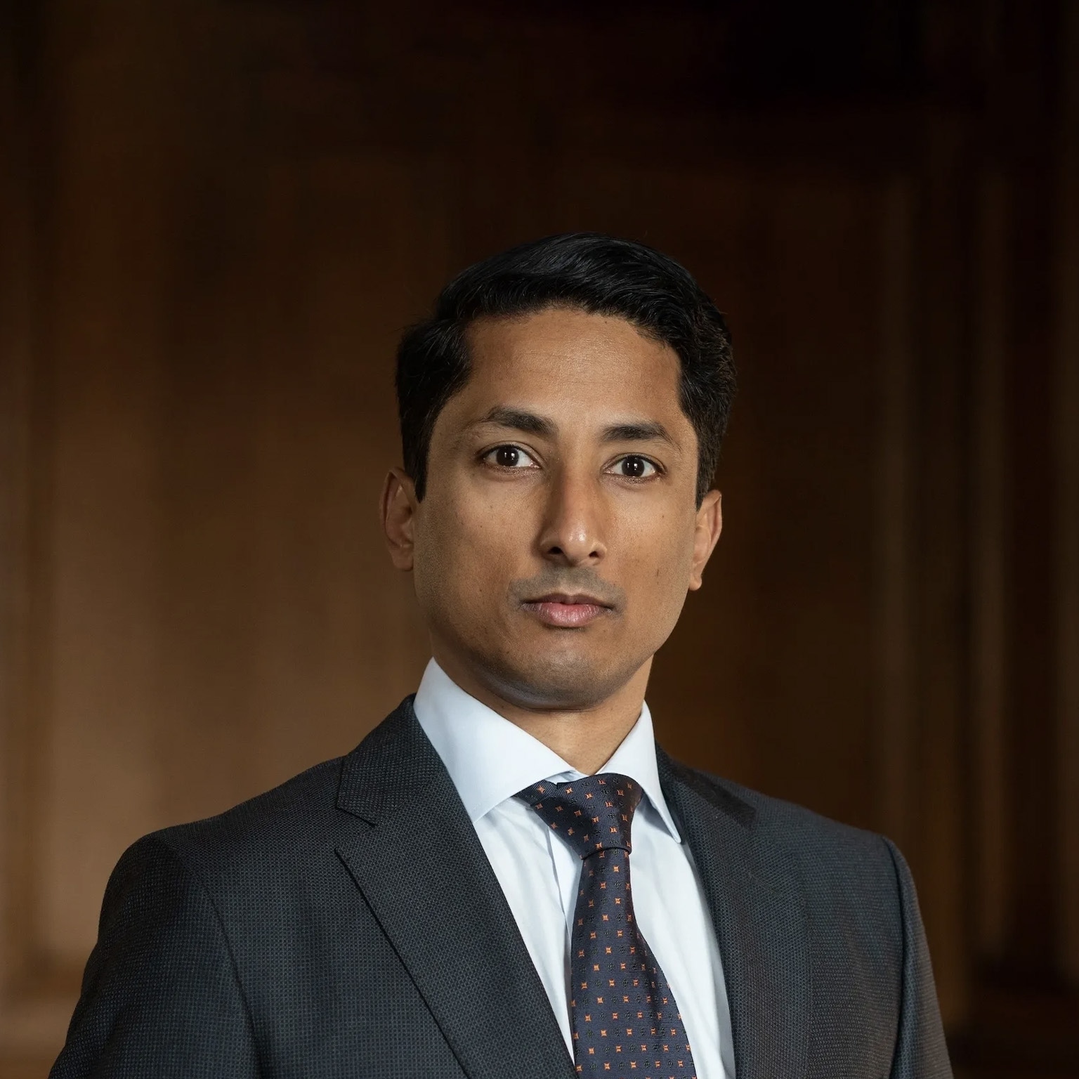 Jean-Paul Carvalho wearing a dark suit, white shirt, and patterned tie looks directly at the camera with a neutral, composed expression. He stands against a warm-toned, softly blurred background. 