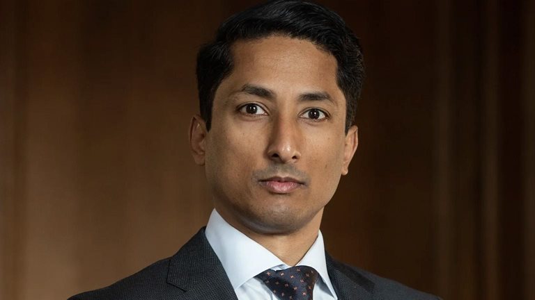 Jean-Paul Carvalh wearing a dark suit, white shirt, and patterned tie looks directly at the camera with a neutral, composed expression. He stands against a warm-toned, softly blurred background. 