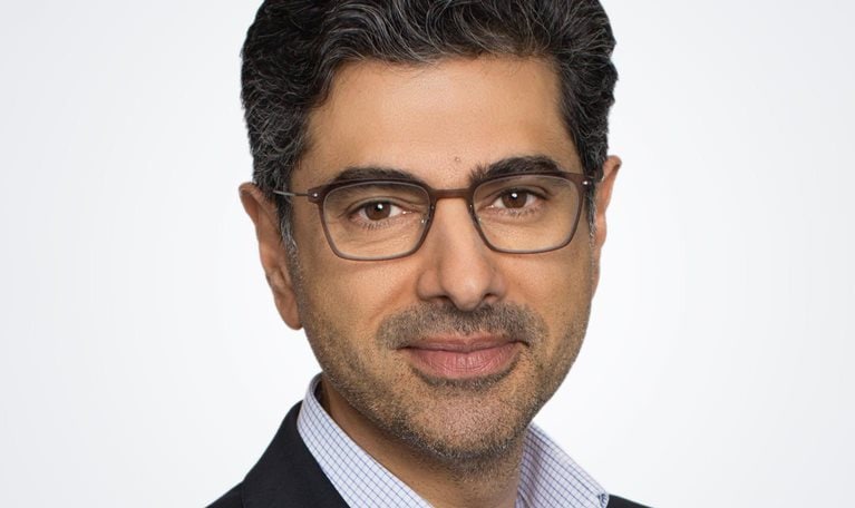 A photograph captures Alok Sama in a suit jacket and button down shirt, smiling gently against a white background.