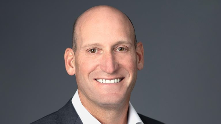David Gitlin is standing against a neutral gray background, dressed in a dark suit jacket and white shirt, with a smile on his face.