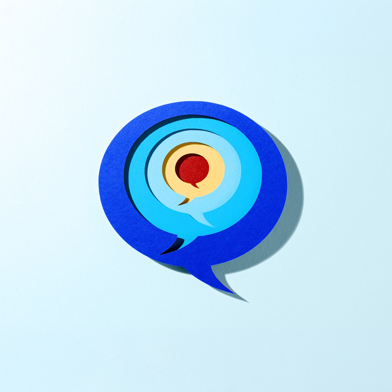 A layered, paper-cut style illustration of overlapping speech bubbles arranged concentrically, with shades of deep blue on the outside and lighter blues toward the center. A yellow and red speech bubble sits at the core, and the entire design casts a soft shadow against a light blue background, creating a subtle three-dimensional effect.