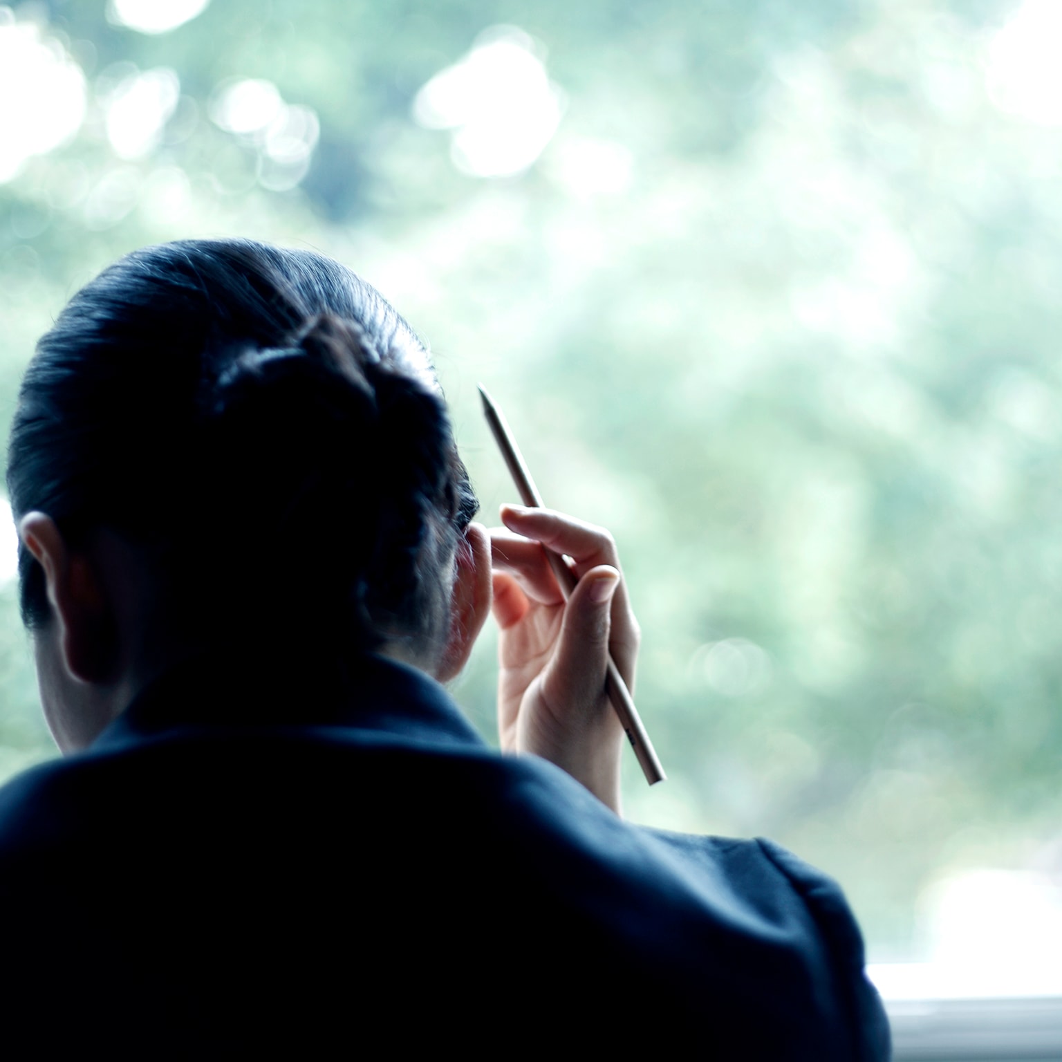 A person in a dark suit, seen from behind, holding a pen and looking out a window toward blurred green foliage.