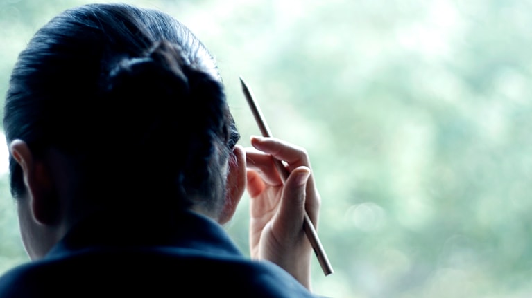 A person in a dark suit, seen from behind, holding a pen and looking out a window toward blurred green foliage.