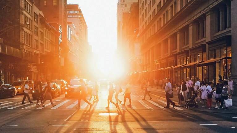 Sunlight shining on the diverse crowds of people walking across the busy intersection on 5th Avenue in New York City.