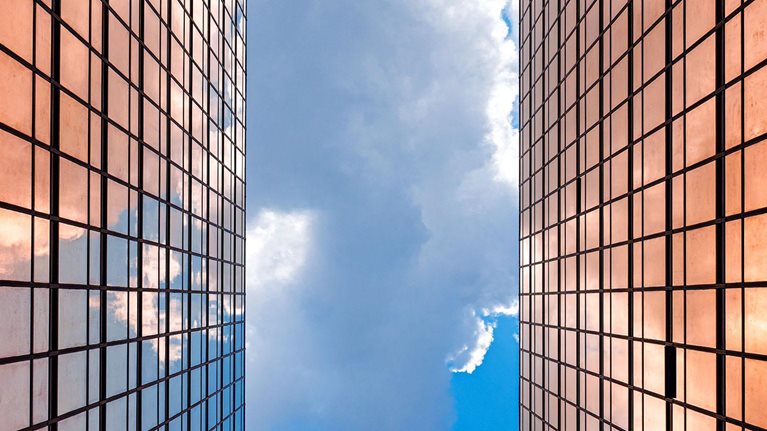 Looking up between two gleaming glass towers toward a vivid blue sky, the city reflects a bright future rising ahead.