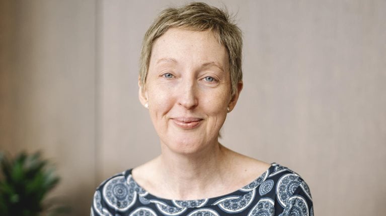 The camera captures CEO Anna Manz's serene smile, framed by her short blonde hair and bright blue eyes. She is wearing a dark blue top with a circular pattern.