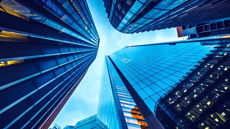 Looking up from street level at the skyscrapers in the heart of London's financial district. The buildings have glass facades and a moody blue cast from the setting sun.