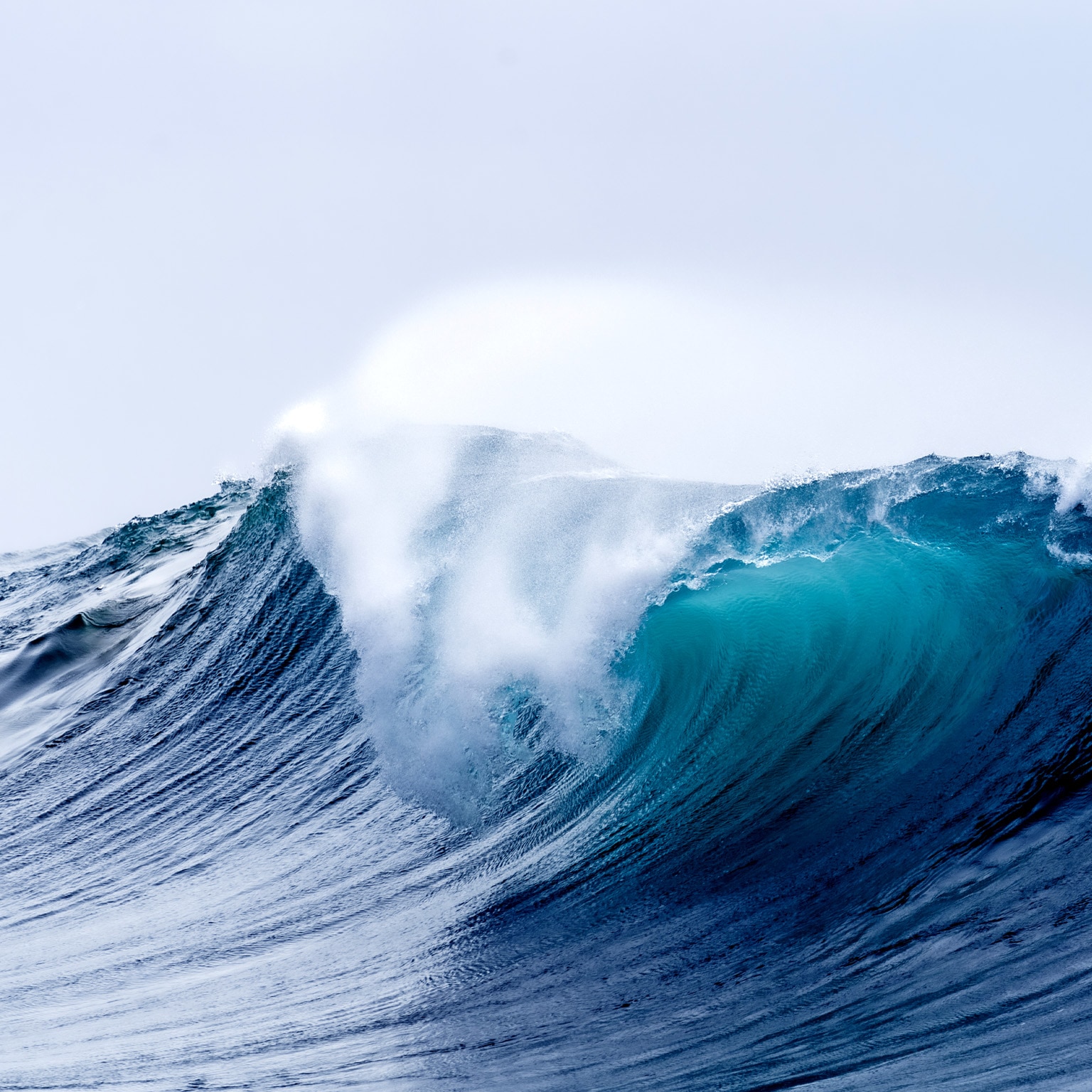 A large ocean wave is captured at its peak, with a deep blue color that transitions to vibrant turquoise as it curls over. The powerful wave is beginning to break, sending a spray of white foam high into the air against a pale sky.
