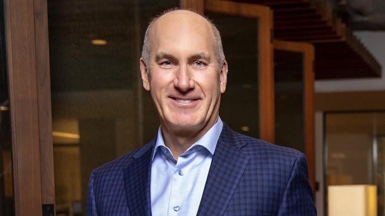 A portrait of John Stankey smiling, wearing a royal blue suit jacket over a blue button-down dress shirt.