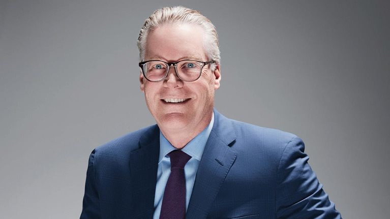 Ed Bastian, sporting a pair of glasses and dressed in a blue suit, light blue shirt, and a purple tie.