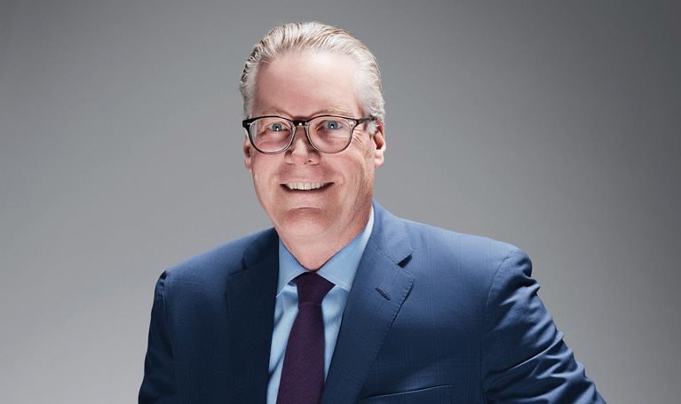 Ed Bastian, sporting a pair of glasses and dressed in a blue suit, light blue shirt, and a purple tie.
