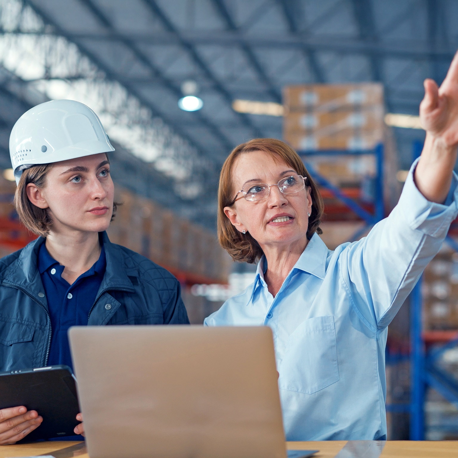  Two women in a warehouse reviewing information on a laptop and tablet, with one wearing a hard hat while the other gestures toward the facility.