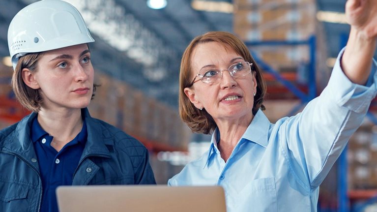 Two women in a warehouse reviewing information on a laptop and tablet, with one wearing a hard hat while the other gestures toward the facility.