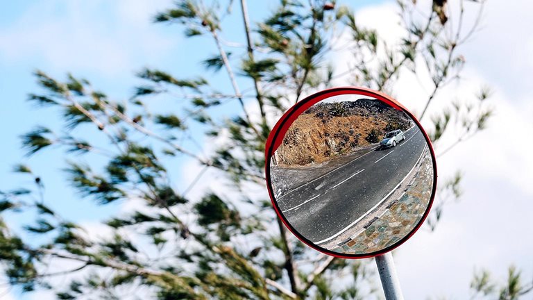 A round roadside convex mirror mounted on a pole, reflecting a winding road with a car driving along it. Behind the mirror, tree branches sway against a bright blue sky with scattered clouds.