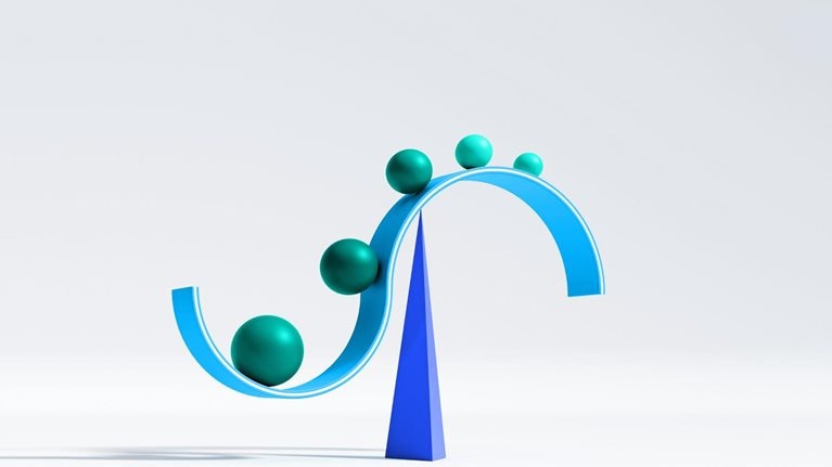 A curved, cyan structure resembling a sideways S is balanced atop a dark blue pyramid. Along the curve of the structure, there are several spheres of varying sizes, starting from the largest and descending in size as they move to a higher point on the curve.