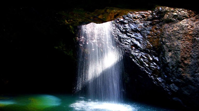  Sunlight streams through a cave opening, illuminating a stunning waterfall plunging into a vibrant turquoise pool below