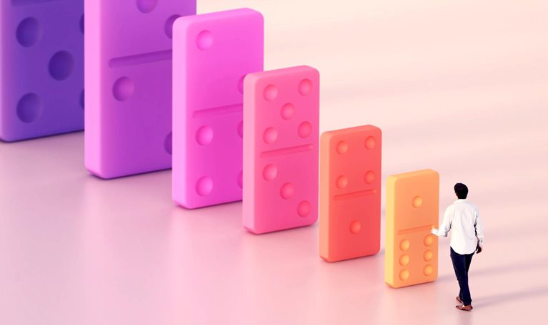 A person approaches a row of increasingly large, colorful dominoes, suggesting a cascading impact.