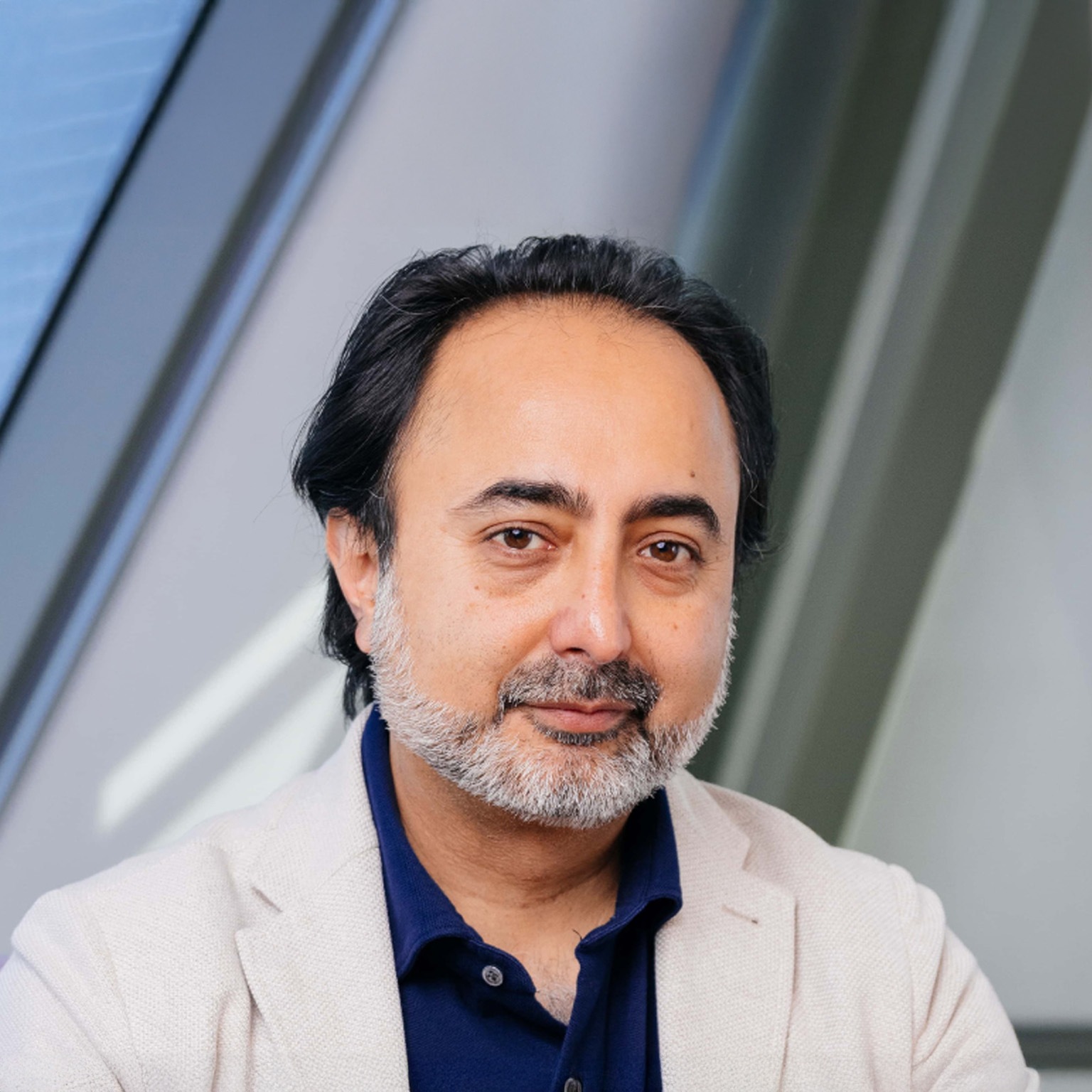 Hemant Taneja with dark hair and a neatly trimmed gray beard looking directly at the camera with a calm, neutral expression. He is wearing a light-colored blazer over a dark blue shirt, standing against a softly blurred indoor background with angled architectural lines.