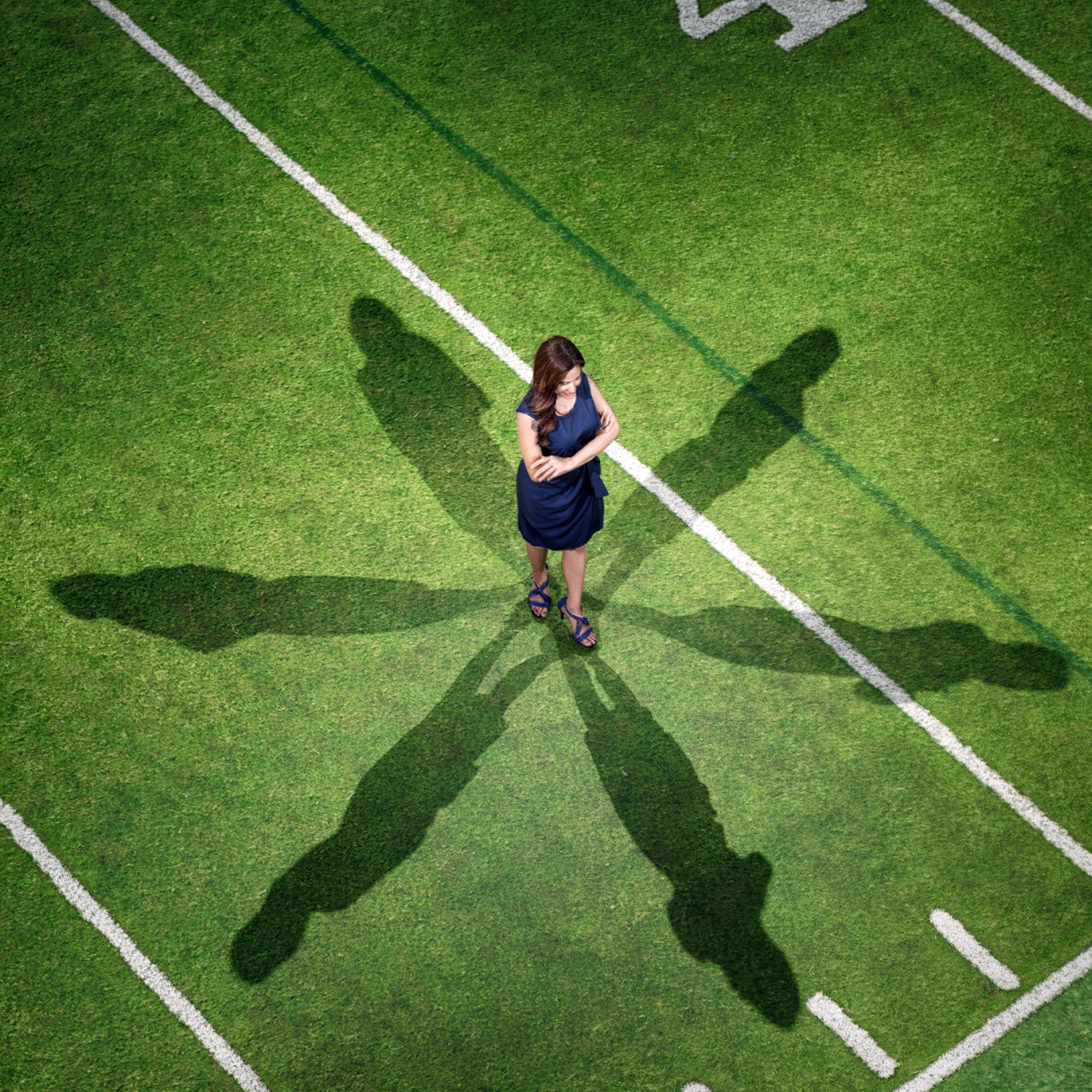 Overhead view of a woman in a dark blue dress standing with her arms crossed on a green sports field, surrounded by six elongated human-shaped shadows. White and dark green field lines cross the grass around her.