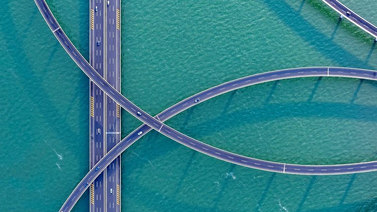 An aerial view of sweeping, elevated highways curving gracefully over turquoise water, creating an intricate, looping pattern. A few cars travel along the roads, adding scale and movement to the striking geometric scene.
