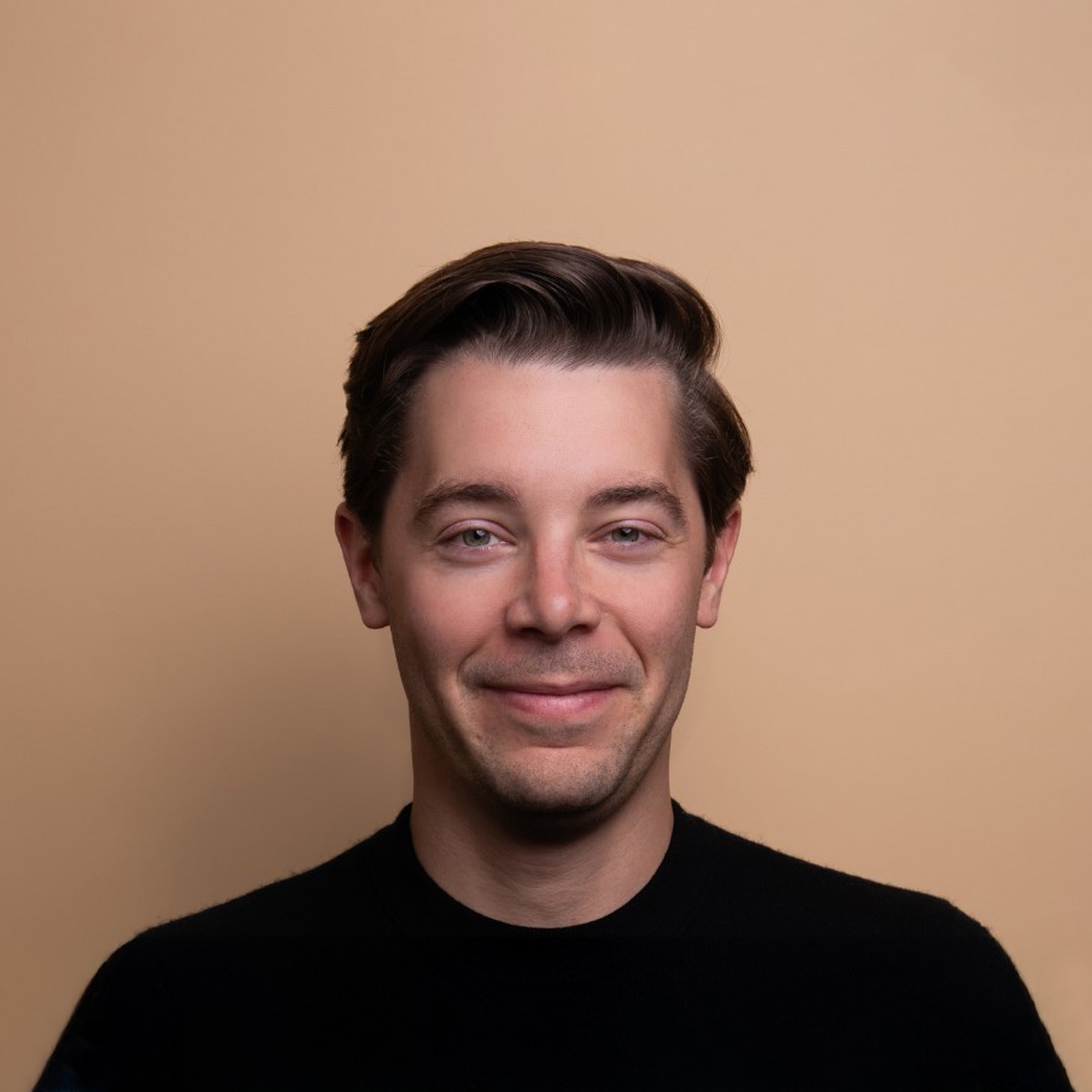 Brad Lightcap with neatly styled dark brown hair looking directly at the camera with a subtle smile. He is wearing a black shirt and is photographed against a plain beige background, creating a simple and professional portrait.