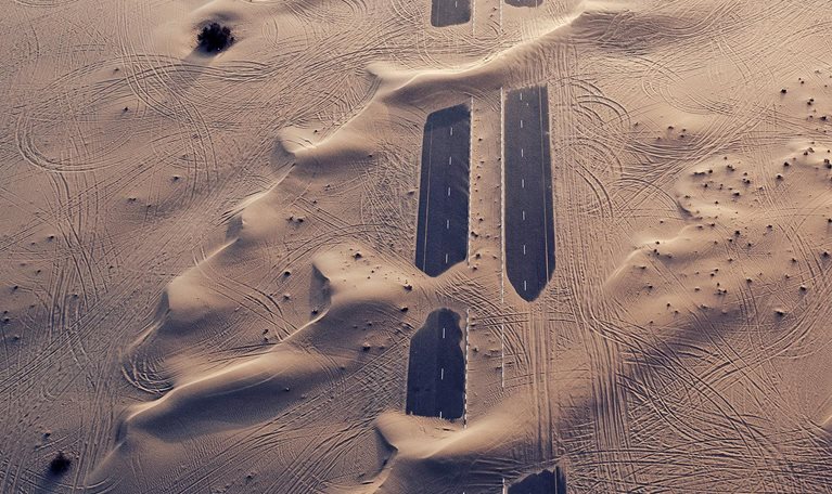 Sandy terrain with a road emerging in parts between stretches of sand.