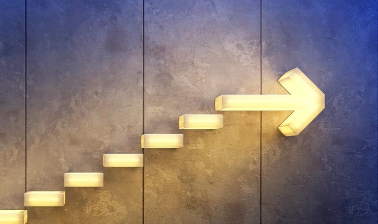 A series of glowing, rectangular stairs arranged in the form of an arrow on a rough concrete surface. The arrow directs upward, indicating progress and accomplishment.