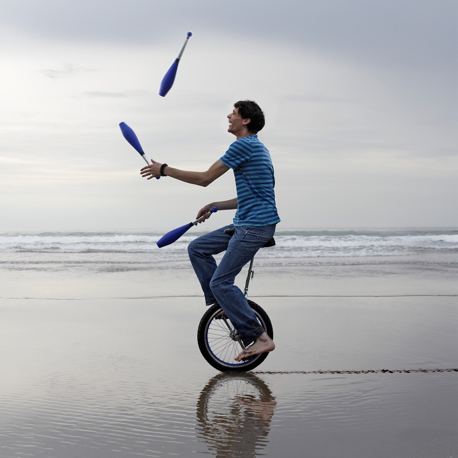 Man sitting on a unicycle while juggling pins at the beach