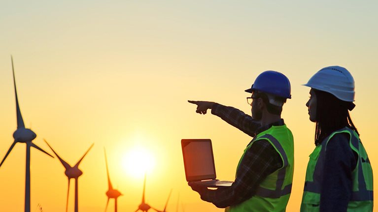 Two workers in a Windmills field working on a laptop and pointing in the direction of the windmills as the sun is setting. 