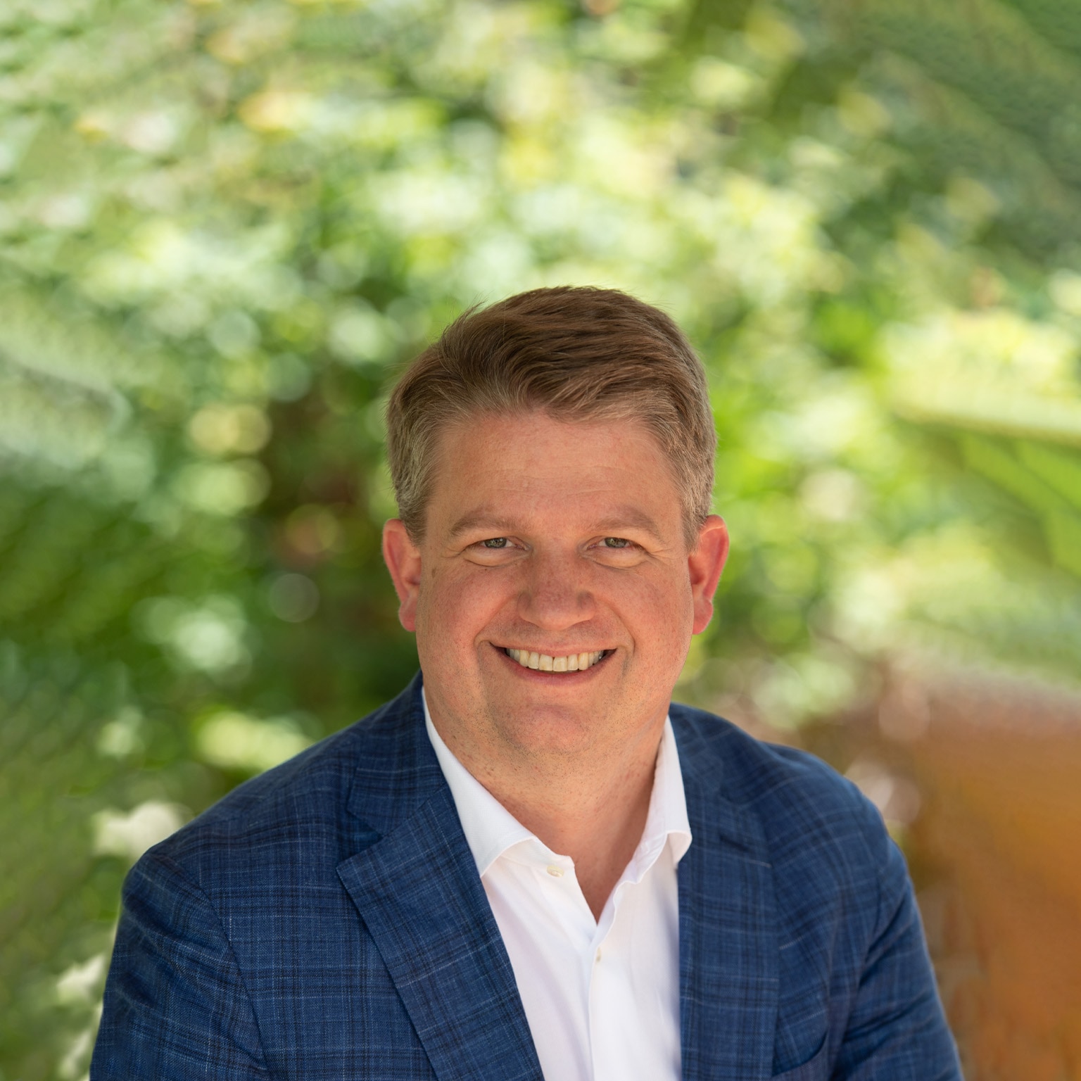 Dave Shull wearing a navy suit jacket and white button-down against a backdrop of lush greenery.