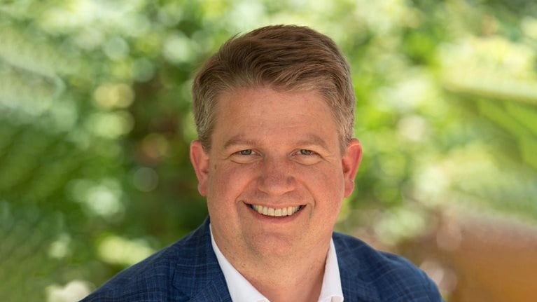 Dave Shull wearing a navy suit jacket and white button-down against a backdrop of lush greenery.