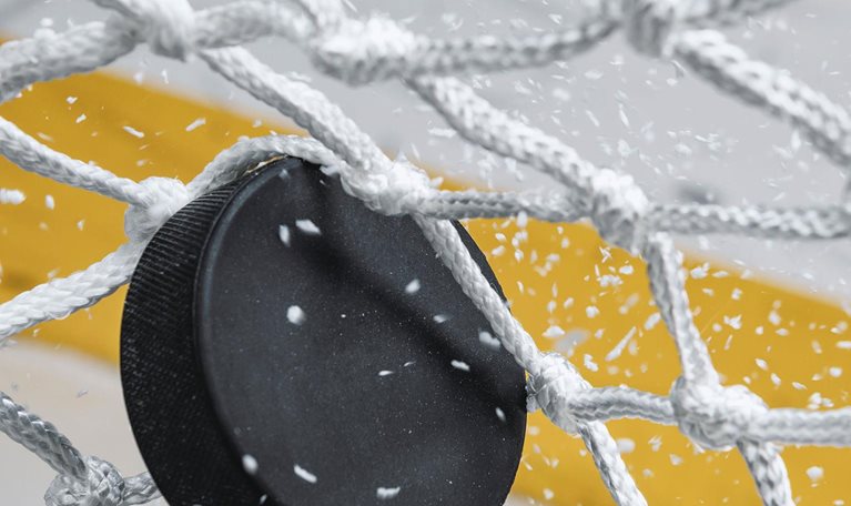 A close-up view of an Ice Hockey puck hitting the back of the goal net as shavings fly by, viewed from the front. Scoring a goal in ice hockey.