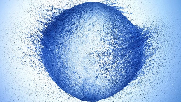 A high-speed photograph of a water balloon bursting. The water is flying outward in all directions, creating a roundish shape with a dynamic and explosive feel.