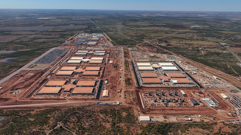 Una foto aérea muestra un centro de datos de Abilene en construcción, con numerosos edificios rectangulares de gran tamaño y extensos movimientos de tierra repartidos en una vasta área. Alrededor del sitio se encuentran campos abiertos, caminos de acceso y grupos de vehículos y equipos, lo que indica un desarrollo activo.