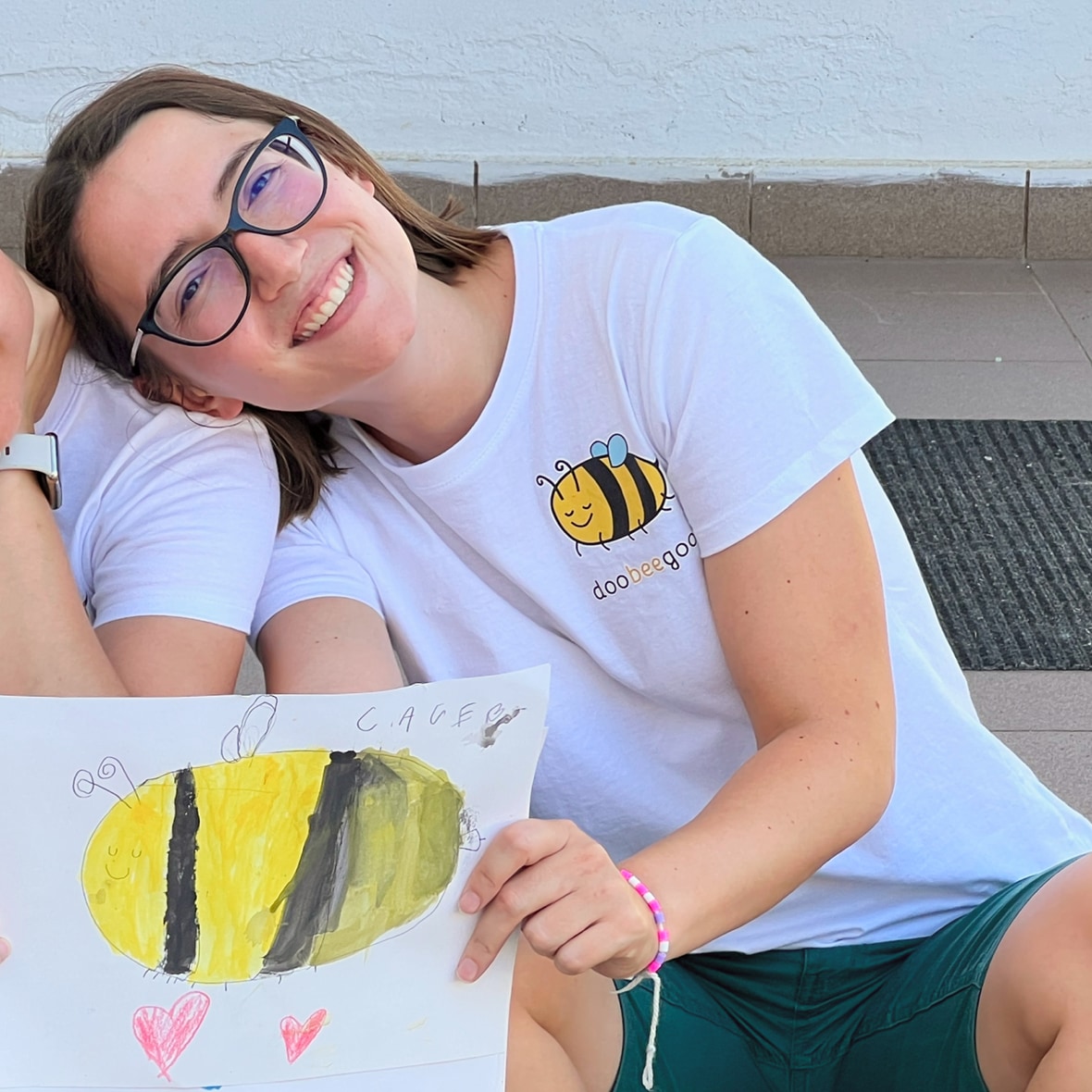 Smiling woman in glasses and a bee t-shirt holds up a child's drawing of a bee.