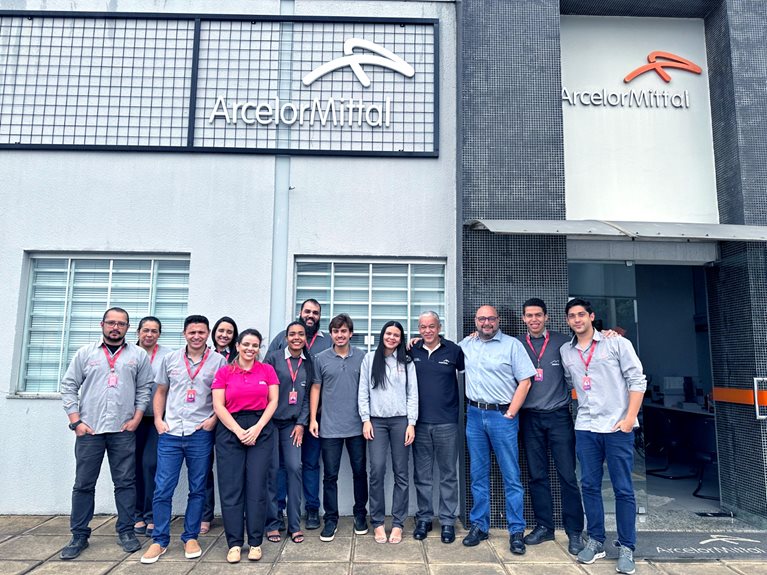 Group of ArcelorMittal employees standing outside an ArcelorMittal facility beneath the company logo.